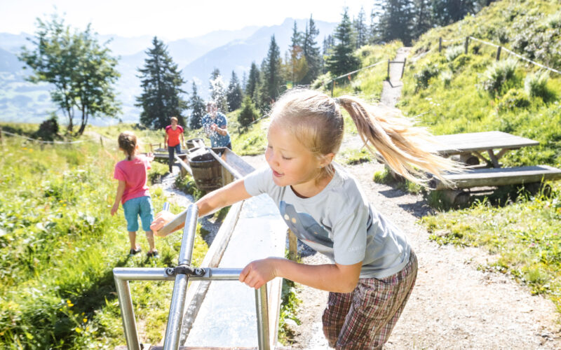 Ein junges Mädchen dreht an einem sonnigen Tag im Freien eine Wasserpumpe mit Metallspirale, während im grasbewachsenen, bergigen Hintergrund Kinder spielen.