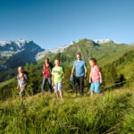 Fünf Personen, darunter Kinder und Erwachsene, wandern auf einem grasbewachsenen Bergpfad mit Bäumen und schneebedeckten Gipfeln im Hintergrund unter einem klaren blauen Himmel.