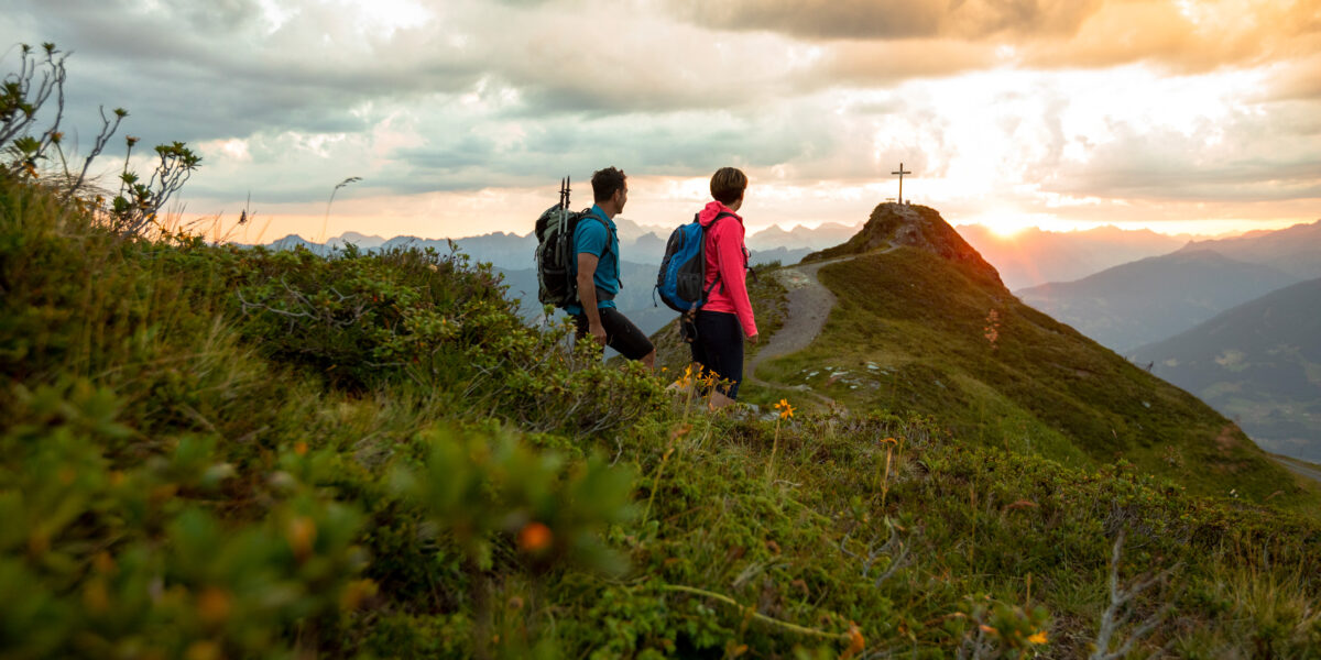 Zwei Wanderer mit Rucksäcken gehen auf einem grasbewachsenen Bergpfad in Richtung eines Gipfels mit einem großen Kreuz bei Sonnenuntergang, umgeben von Wolken und fernen Gipfeln.