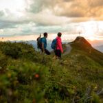 Zwei Wanderer mit Rucksäcken gehen auf einem grasbewachsenen Bergpfad in Richtung eines Gipfels mit einem großen Kreuz bei Sonnenuntergang, umgeben von Wolken und fernen Gipfeln.