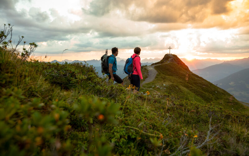 Zwei Wanderer mit Rucksäcken gehen auf einem grasbewachsenen Bergpfad in Richtung eines Gipfels mit einem großen Kreuz bei Sonnenuntergang, umgeben von Wolken und fernen Gipfeln.
