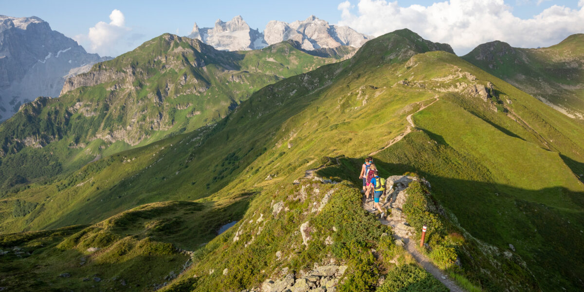 Eine Gruppe von Wanderern wandert auf einem schmalen Bergpfad mit grünen Hügeln und fernen felsigen Gipfeln unter einem teilweise bewölkten Himmel.