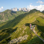 Eine Gruppe von Wanderern wandert auf einem schmalen Bergpfad mit grünen Hügeln und fernen felsigen Gipfeln unter einem teilweise bewölkten Himmel.