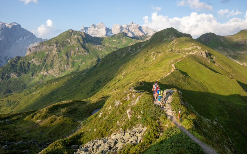 Eine Gruppe von Wanderern wandert auf einem schmalen Bergpfad mit grünen Hügeln und fernen felsigen Gipfeln unter einem teilweise bewölkten Himmel.