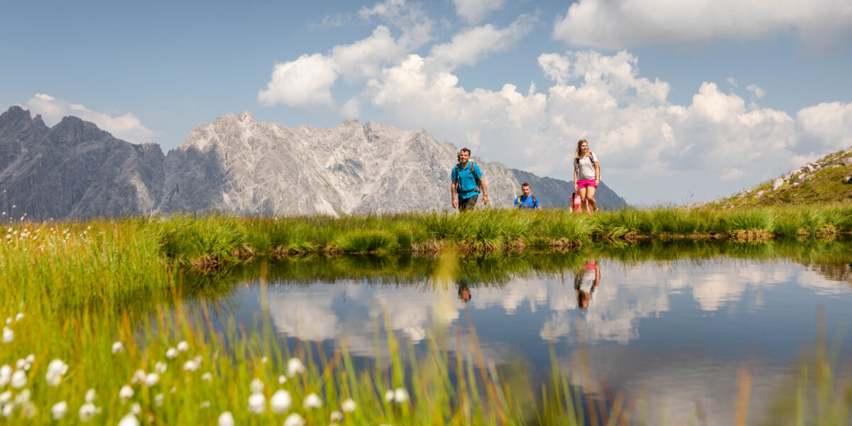 Vier Personen wandern an einem ruhigen Bergsee, mit hohem Gras und weißen Wildblumen im Vordergrund und schroffen Gipfeln unter einem teilweise bewölkten Himmel im Hintergrund.