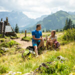 Zwei Personen sitzen auf einem grasbewachsenen Bergpfad, unterhalten sich und essen eine Kleinigkeit, mit Rucksäcken in der Nähe und einer alpinen Landschaft im Hintergrund.
