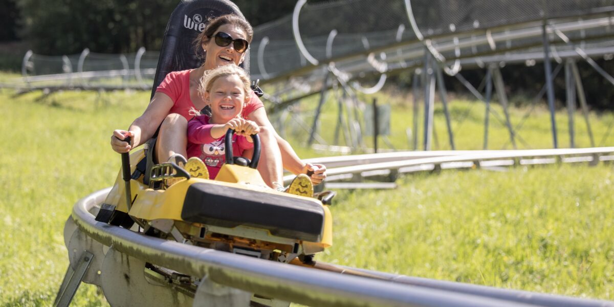 Ein Erwachsener und ein Kind fahren zusammen in einem gelben Wagen auf einer Outdoor-Alpine-Coaster-Bahn, umgeben von grünem Gras und Bäumen.
