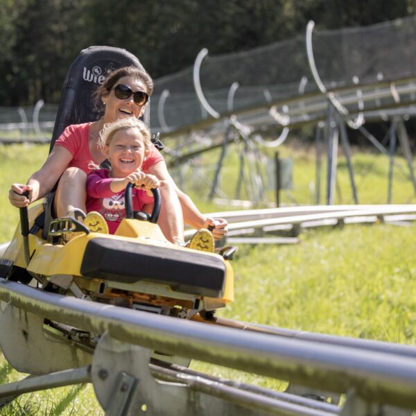 Ein Erwachsener und ein Kind fahren zusammen in einem gelben Wagen auf einer Outdoor-Alpine-Coaster-Bahn, umgeben von grünem Gras und Bäumen.