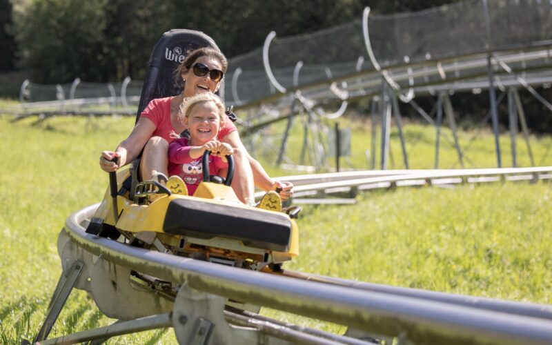 Ein Erwachsener und ein Kind fahren zusammen in einem gelben Wagen auf einer Outdoor-Alpine-Coaster-Bahn, umgeben von grünem Gras und Bäumen.