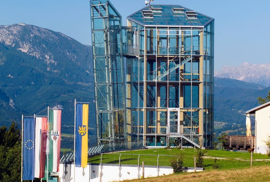 Ein modernes Glasgebäude mit Außentreppen steht auf einem grasbewachsenen Hügel, vor dem mehrere Fahnen wehen und im Hintergrund Berge zu sehen sind.