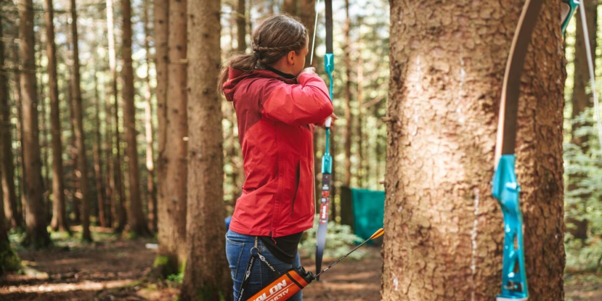 Eine Person in einer roten Jacke zielt mit Pfeil und Bogen in einem Waldstück, im Vordergrund lehnt ein weiterer Bogen an einem Baum.