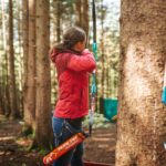 Eine Person in einer roten Jacke zielt mit Pfeil und Bogen in einem Waldstück, im Vordergrund lehnt ein weiterer Bogen an einem Baum.