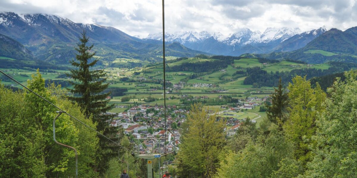 Menschen fahren mit einem Sessellift über ein grünes Tal mit einem Dorf darunter, umgeben von Bäumen und schneebedeckten Bergen im Hintergrund unter einem teilweise bewölkten Himmel.