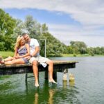 Ein Paar sitzt zusammen auf einem hölzernen Steg mit den Füßen im Wasser, umgeben von Bäumen und Grünpflanzen unter einem teilweise bewölkten Himmel.