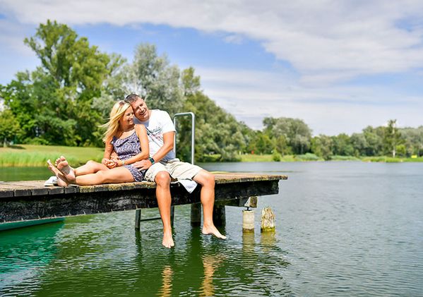 Ein Paar sitzt zusammen auf einem hölzernen Steg mit den Füßen im Wasser, umgeben von Bäumen und Grünpflanzen unter einem teilweise bewölkten Himmel.