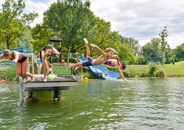 Vier Kinder springen von einem Steg in einen See, im Hintergrund sind grüne Bäume und eine Wasserrutsche zu sehen.