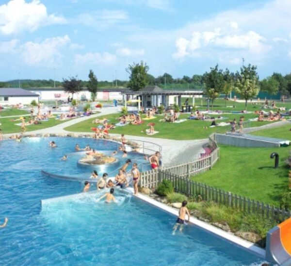 Öffentliches Freibad mit Menschen, die schwimmen, sich auf dem Rasen entspannen und an einem sonnigen Tag eine große orangefarbene Wasserrutsche benutzen.