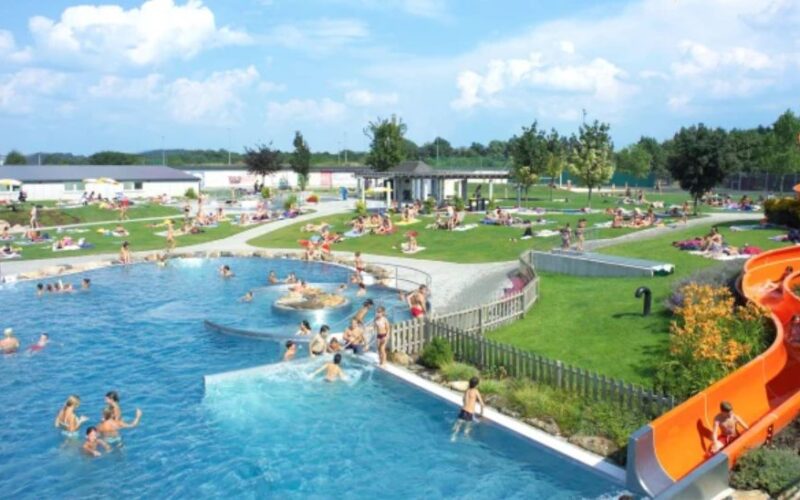 Öffentliches Freibad mit Menschen, die schwimmen, sich auf dem Rasen entspannen und an einem sonnigen Tag eine große orangefarbene Wasserrutsche benutzen.