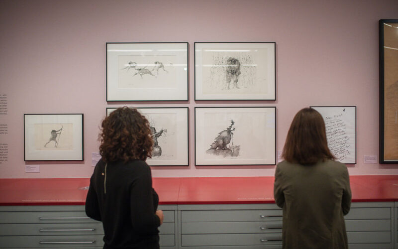 Zwei Personen betrachten gerahmte Tierskizzen, die in einer Galerie oder einem Museum an einer rosafarbenen Wand über einer roten Theke ausgestellt sind.