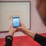 Eine Person macht mit einem Smartphone ein Foto von einem gerahmten Kunstwerk an der Wand. Das Bild auf dem Bildschirm des Telefons entspricht dem fotografierten Kunstwerk.