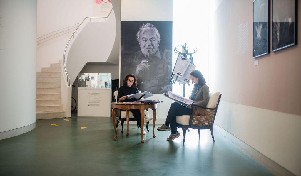 Zwei Personen sitzen an einem Tisch und lesen Dokumente in einem modernen Galerieraum, in dem ein großes Schwarz-Weiß-Porträt und Fotos an den Wänden hängen.