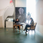 Zwei Personen sitzen an einem Tisch und lesen Dokumente in einem modernen Galerieraum, in dem ein großes Schwarz-Weiß-Porträt und Fotos an den Wänden hängen.