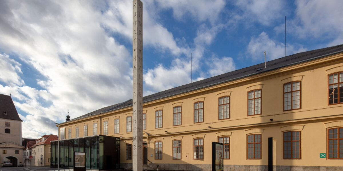 Ein großes beigefarbenes Gebäude mit gewölbten Fenstern und einem hohen vertikalen Schild mit der Aufschrift "KUNSTHALLE" unter einem teilweise bewölkten Himmel.
