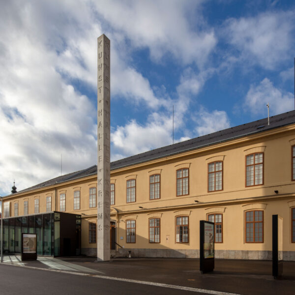 Ein großes beigefarbenes Gebäude mit gewölbten Fenstern und einem hohen vertikalen Schild mit der Aufschrift "KUNSTHALLE" unter einem teilweise bewölkten Himmel.