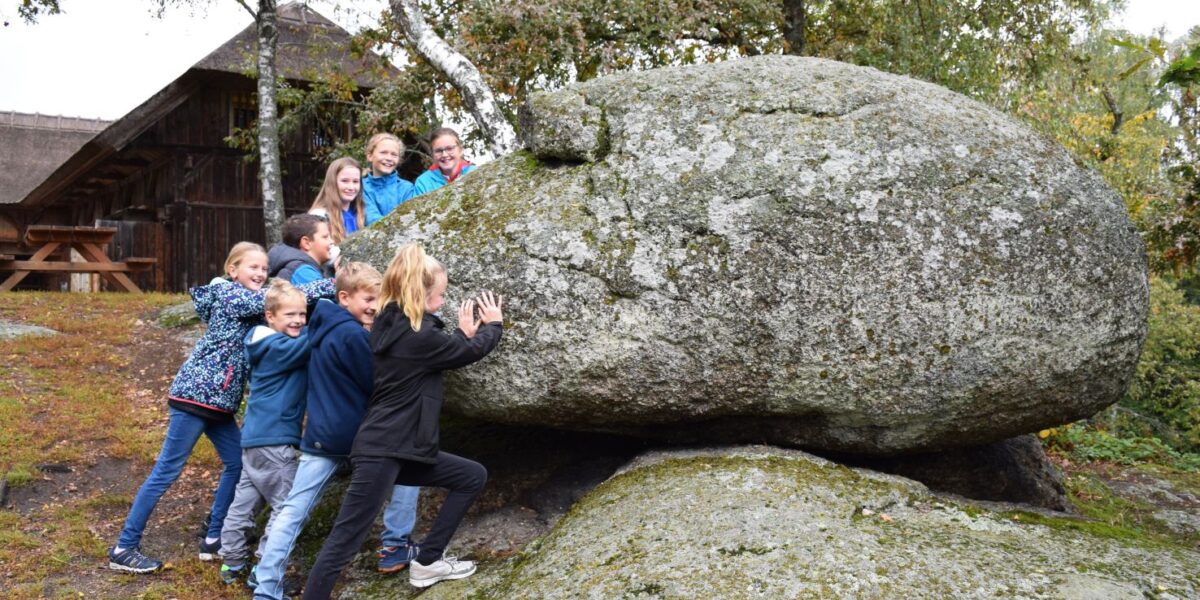 Eine Gruppe Kinder stemmt sich im Freien gegen einen großen, moosbedeckten Felsbrocken, im Hintergrund sind Bäume und ein Holzgebäude zu sehen.
