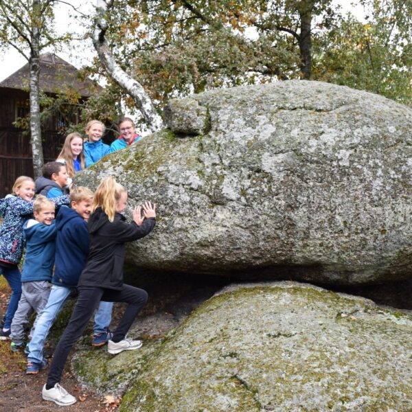 Eine Gruppe Kinder stemmt sich im Freien gegen einen großen, moosbedeckten Felsbrocken, im Hintergrund sind Bäume und ein Holzgebäude zu sehen.