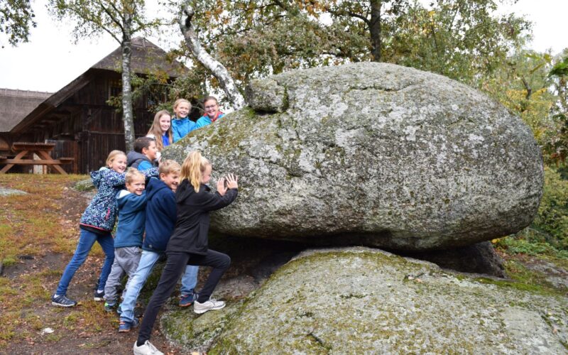 Eine Gruppe Kinder stemmt sich im Freien gegen einen großen, moosbedeckten Felsbrocken, im Hintergrund sind Bäume und ein Holzgebäude zu sehen.
