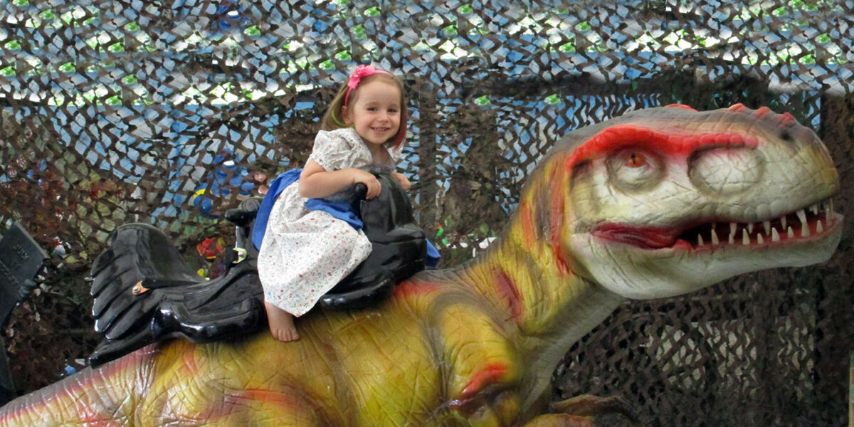 Ein junges Mädchen in einem weißen Kleid mit rosa Blumen sitzt auf einer großen Dinosaurier-Statue und lächelt in die Kamera. Im Hintergrund ist ein Tarnnetz zu sehen.