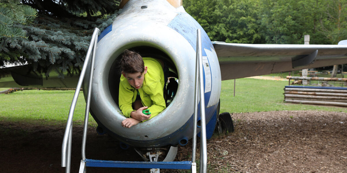 Eine Person in einer grünen Jacke liegt in der vorderen Öffnung eines stationären Düsenflugzeugs, das im Freien in der Nähe von Bäumen und Gras ausgestellt ist.