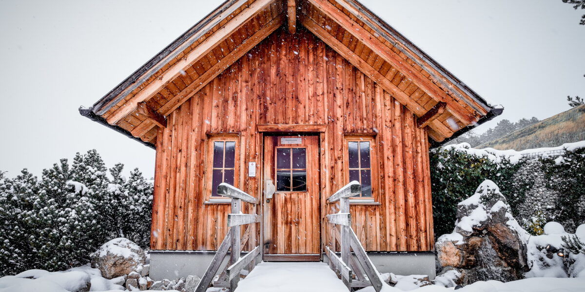 Eine kleine Holzhütte mit steilem Dach steht in einer verschneiten Landschaft, flankiert von Bäumen und Felsen, unter fallendem Schnee.