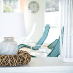 Eine weiße Lampe mit einem Korbsockel steht auf einer weißen Fläche; im Hintergrund stehen tealfarbene Loungesessel, eine Uhr und helle Fenster.