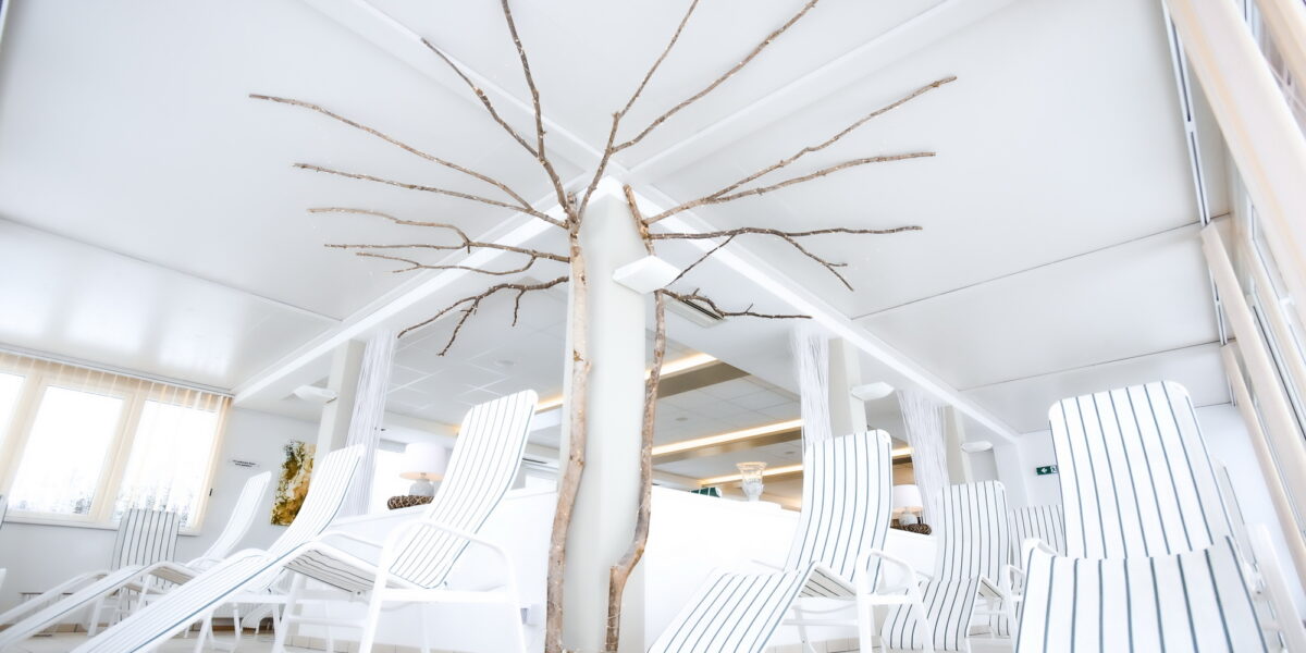 Ein blattloser Baum, umgeben von weißen Lounge-Sesseln mit schwarzen Streifen, in einem hellen, modernen Innenraum mit großen Fenstern.