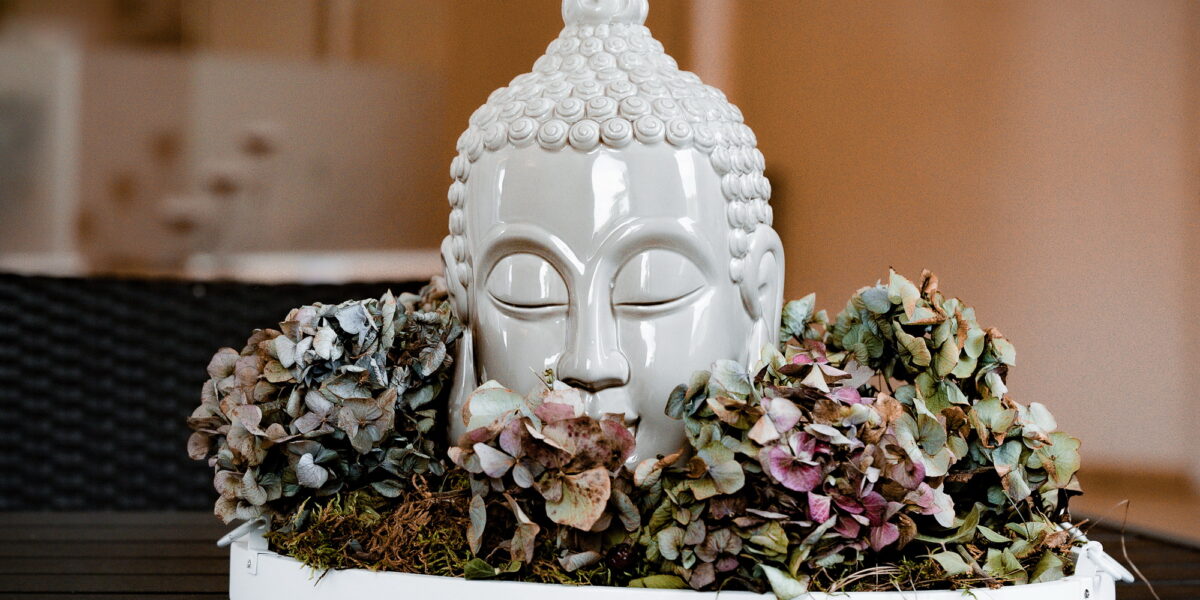 Ein weißer Keramik-Buddha-Kopf ist von getrockneten Hortensienblüten und Moos umgeben und steht auf einem runden Tablett auf einem dunklen Tisch.