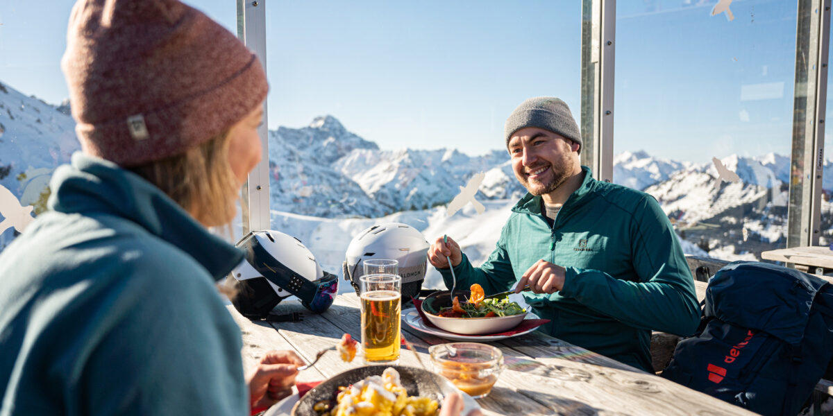 Zwei Personen in Winterkleidung essen an einem Tisch im Freien mit Blick auf die Berge zu Mittag, die Helme liegen auf dem Tisch und die Getränke neben den Tellern.