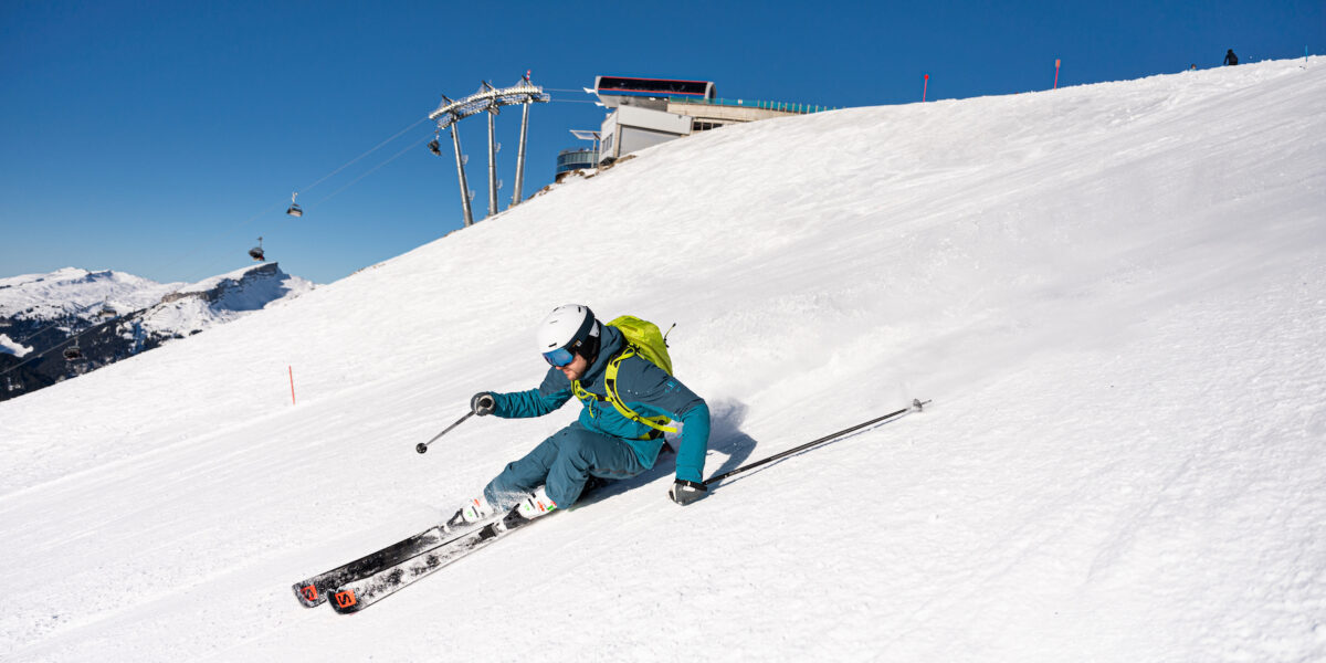 Ein Skifahrer im blauen Outfit und mit Helm fährt an einem klaren, sonnigen Tag einen verschneiten Hang in der Nähe eines Skilifts hinunter.