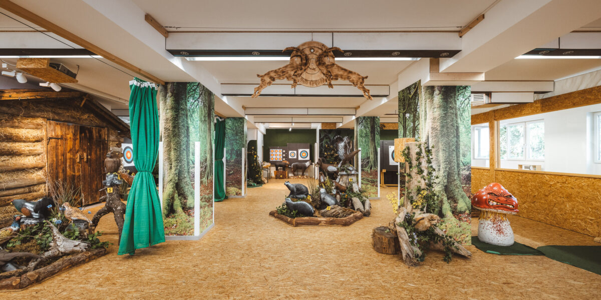 Indoor-Bogenschießanlage mit waldähnlichem Dekor, Tierzielen, Bogenschießzielen im hinteren Bereich und Naturholzböden bei heller Beleuchtung.
