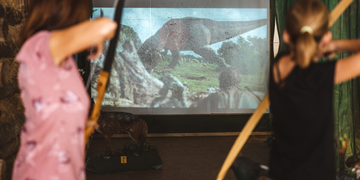 Zwei Personen üben sich in einem Raum im Bogenschießen und zielen auf einen Bildschirm, der eine Dinosaurierszene aus einem Film oder Spiel zeigt.
