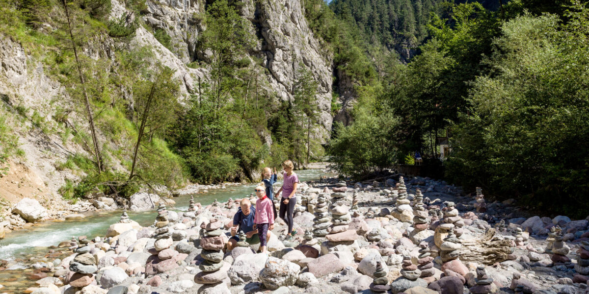 Menschen stehen zwischen zahlreichen aufgeschichteten Steinhaufen an einem felsigen Flussufer, umgeben von Bäumen und steilen Klippen unter einem klaren blauen Himmel.