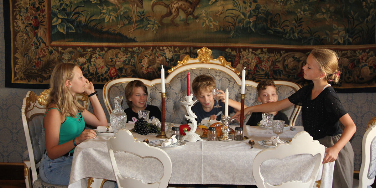 Fünf Kinder sitzen um einen eleganten Esstisch mit Tee und Essen in einem Raum, der mit einem kunstvollen Wandteppich und alten Möbeln geschmückt ist.