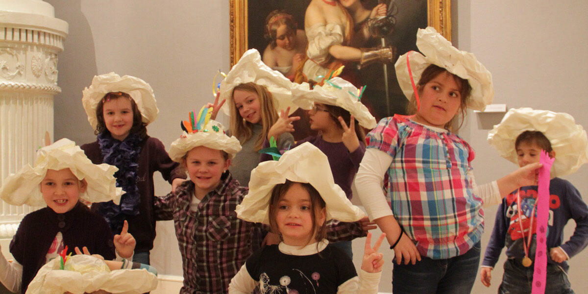 Eine Gruppe von Kindern mit großen Papierhüten posiert vor einem klassischen Gemälde in einem Kunstmuseum.
