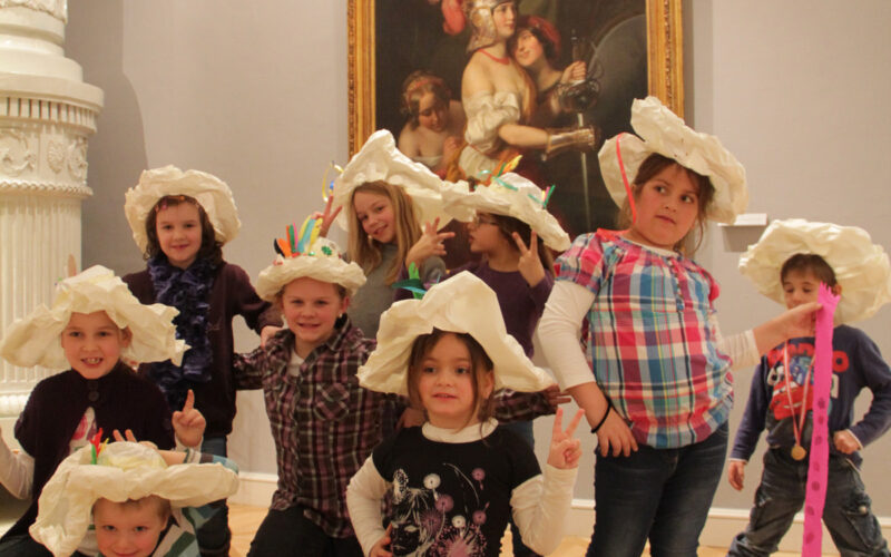 Eine Gruppe von Kindern mit großen Papierhüten posiert vor einem klassischen Gemälde in einem Kunstmuseum.