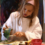 Ein junges Mädchen in einem weißen Kittel malt mit einem Pinsel an einem Tisch, umgeben von bunten Schalen und Kunstzubehör, mit abstrakten Kunstwerken im Hintergrund.