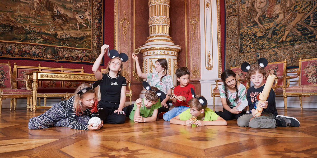 Eine Gruppe von Kindern mit Mausohr-Stirnbändern spielt auf dem verzierten Holzfußboden eines prächtigen, mit Gobelins geschmückten Saals.