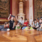 Eine Gruppe von Kindern mit Mausohr-Stirnbändern spielt auf dem verzierten Holzfußboden eines prächtigen, mit Gobelins geschmückten Saals.