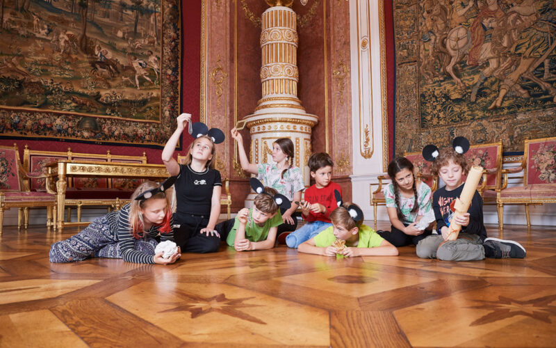 Eine Gruppe von Kindern mit Mausohr-Stirnbändern spielt auf dem verzierten Holzfußboden eines prächtigen, mit Gobelins geschmückten Saals.