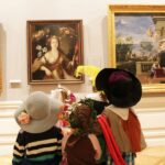 Eine Gruppe von Kindern in Kostümen steht vor gerahmten Gemälden in einem Kunstmuseum und betrachtet die Kunstwerke.